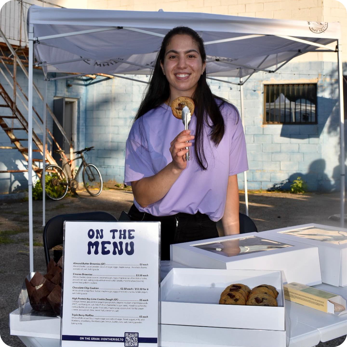 The On the Rise owner Zaina at a local market in her stall holding a cookie with a pair of tongs.